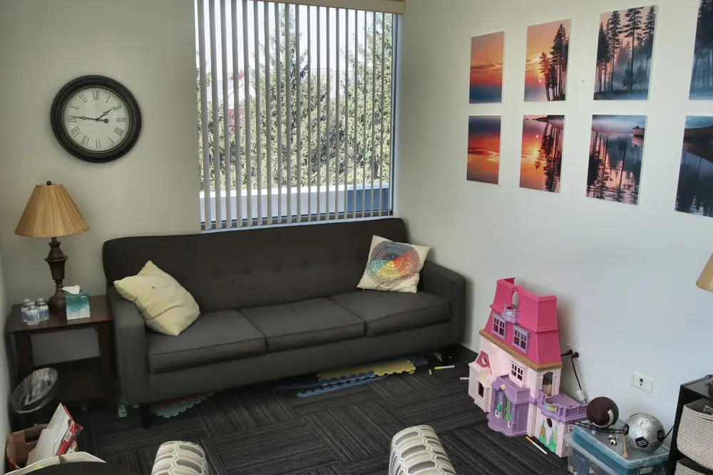 A counseling room at the Geneva office, with a dark sofa and bright pillows, a wall clock, colorful framed art, and a window with blinds.