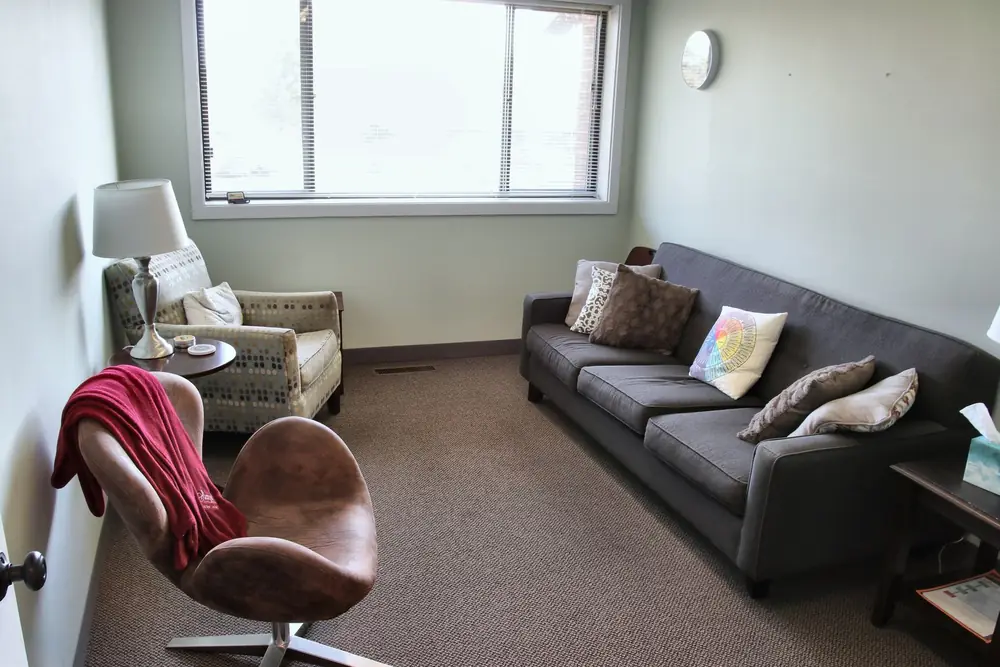 A counseling room at the Naperville office, with a gray sofa and pillows, a brown swivel chair with a red throw, and a patterned armchair by the window.