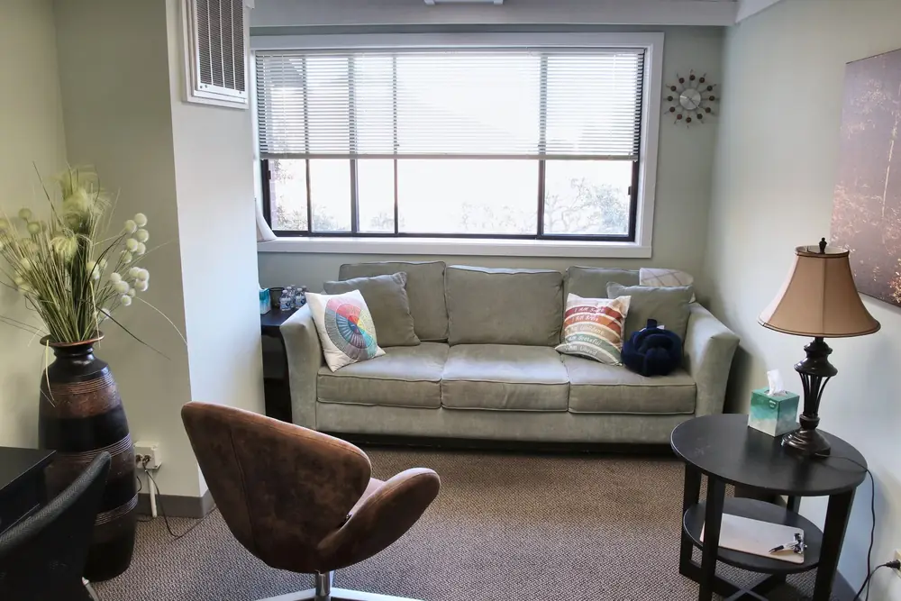 A counseling room at the Naperville office, with a sage loveseat, a brown swivel chair, a table lamp, and a tall vase of flowers.