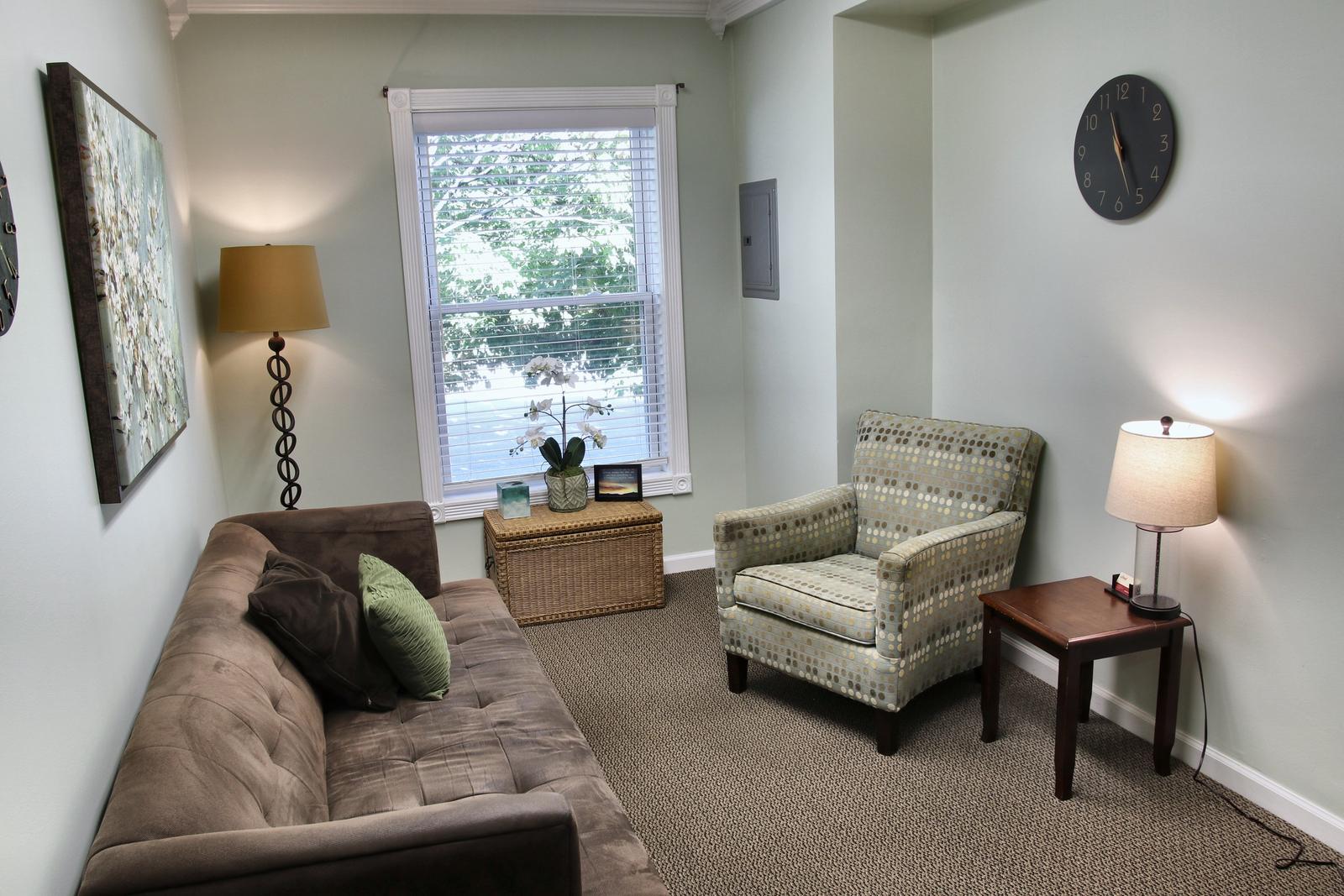 A counseling room at the Wheaton office, with a brown sofa, a patterned armchair, a lamp on a side table, and a window looking out on trees.